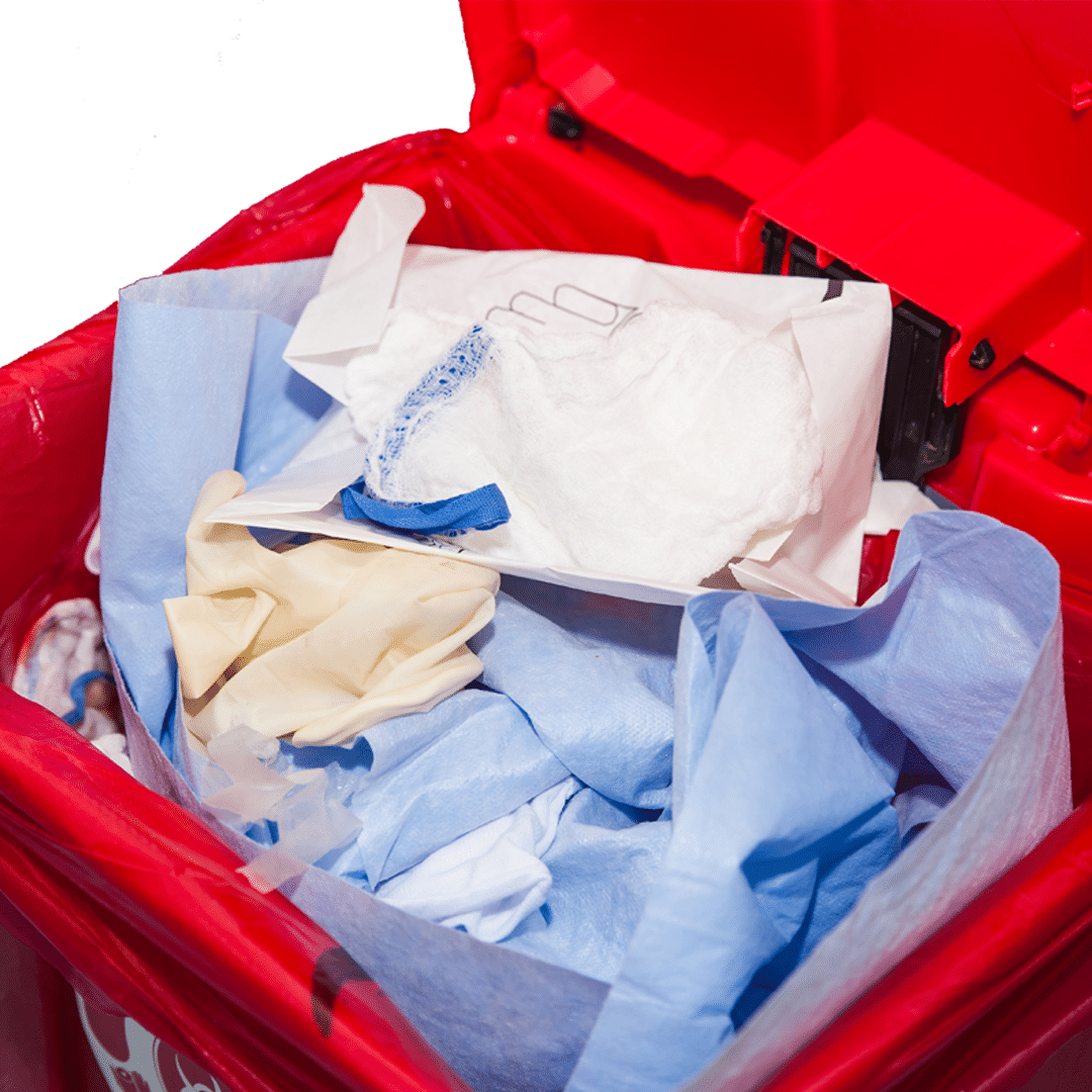 içerisinde mavi ameliyat önlüğü beyaz eldiven tek kullanımlık maske bulununan bir hastanenin ameliyathanesinde bulunan kırmızı çöp kutusu
